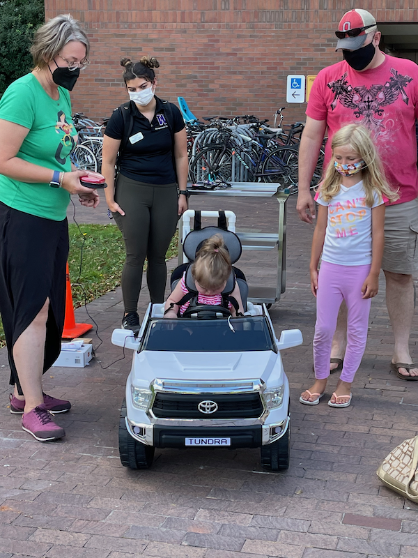 a group of workshop participants and parents testing out a switch during a workshop, with a young girl with blond hair and a pink shirt sitting in a white Go Baby Go car