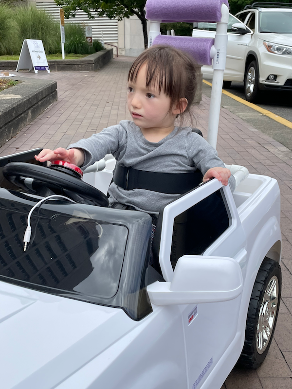 a young girl with brown hair and wearing a gray shirt is sitting in a white Go Baby Go car and pushing her red 'Go' button