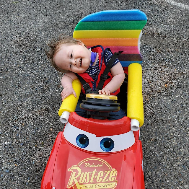A young boy with brown hair and waring a blue and red shirt leans over the rail of his Go Baby Go car, smiling and pushing his yellow 'Go' button