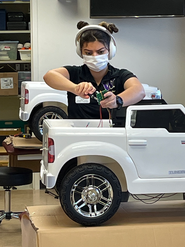 a woman with dark brown hair wearing headphones, a white mask, and black shirt, using tools to modify a white Go Baby Go car during a workshop.