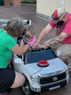 two adults kneeling down next to a Go Baby Go car with a red switch on the car hood, making adjustments for a young girl with blond hair wearing a pink shirt.