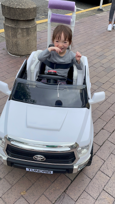 a young girl with brown hair and a gray shirt is sitting in a white Go Baby Go car. She is smiling, with her hands up to her mouth..