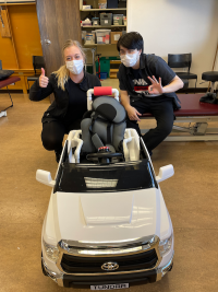 two adult volunteers, both dressed in black and wearing masks, kneel next to a completed Go Baby Go car they have just modified, giving the thumbs up sign.