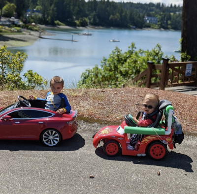 Two brothers driving their Go Baby Go cars together on a sidewalk, with trees and water behind them in the distance.