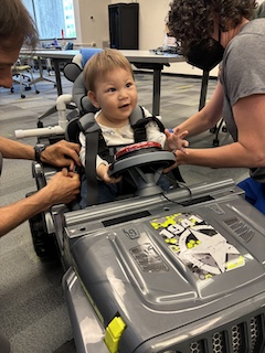 A boy with a long sleeved shirt and short brown hair sits in his Go Baby Go car, looking at an adult and reaching for his big red button