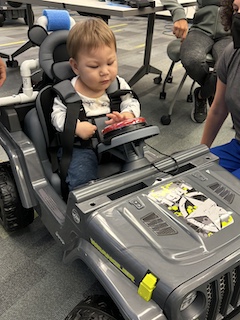 A boy with a long sleeved shirt and short brown hair sits in his Go Baby Go car, pressing his big red button