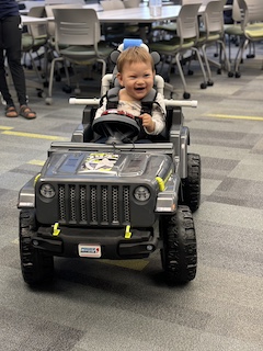 A boy with a long sleeved shirt and short brown hair sits in his Go Baby Go car, laughing