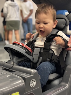 A boy with a long sleeved shirt and short brown hair sits in his Go Baby Go car, looking at his big red button with his mouth wide open