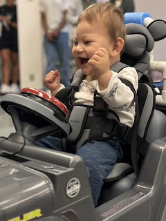 A boy with a long sleeved shirt and short brown hair sits in his Go Baby Go car, looking at his big red button and laughing