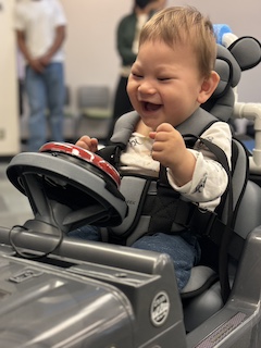 A boy with a long sleeved shirt and short brown hair sits in his Go Baby Go car, looking at his big red button and laughing