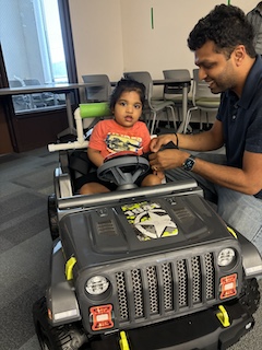 A boy with an orange shirt and long dark hair in in a bun sits in his Go Baby Go car, at the camera while his dad helps him reach for his big red button