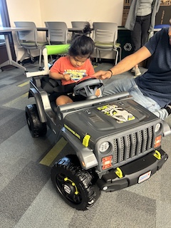 A boy with an orange shirt and long dark hair up in a bun sits in his Go Baby Go car, looking down as his dad presses his big red button