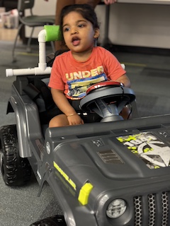 A boy with an orange shirt and long dark hair up in a bun sits in his Go Baby Go car, smiling with an open mouth and reaching for his button