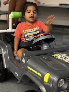 A boy with an orange shirt and long dark hair up in a bun sits in his Go Baby Go car, smiling and reaching for his button