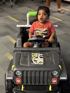 A boy with an orange shirt and long dark hair in in a bun sits in his Go Baby Go car, smiling softly at the camera