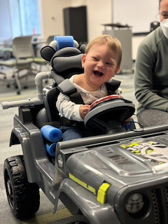 A boy with a long sleeved shirt and short brown hair sits in his Go Baby Go car, laughing