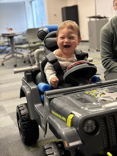 A boy with a long sleeved shirt and short brown hair sits in his Go Baby Go car, laughing